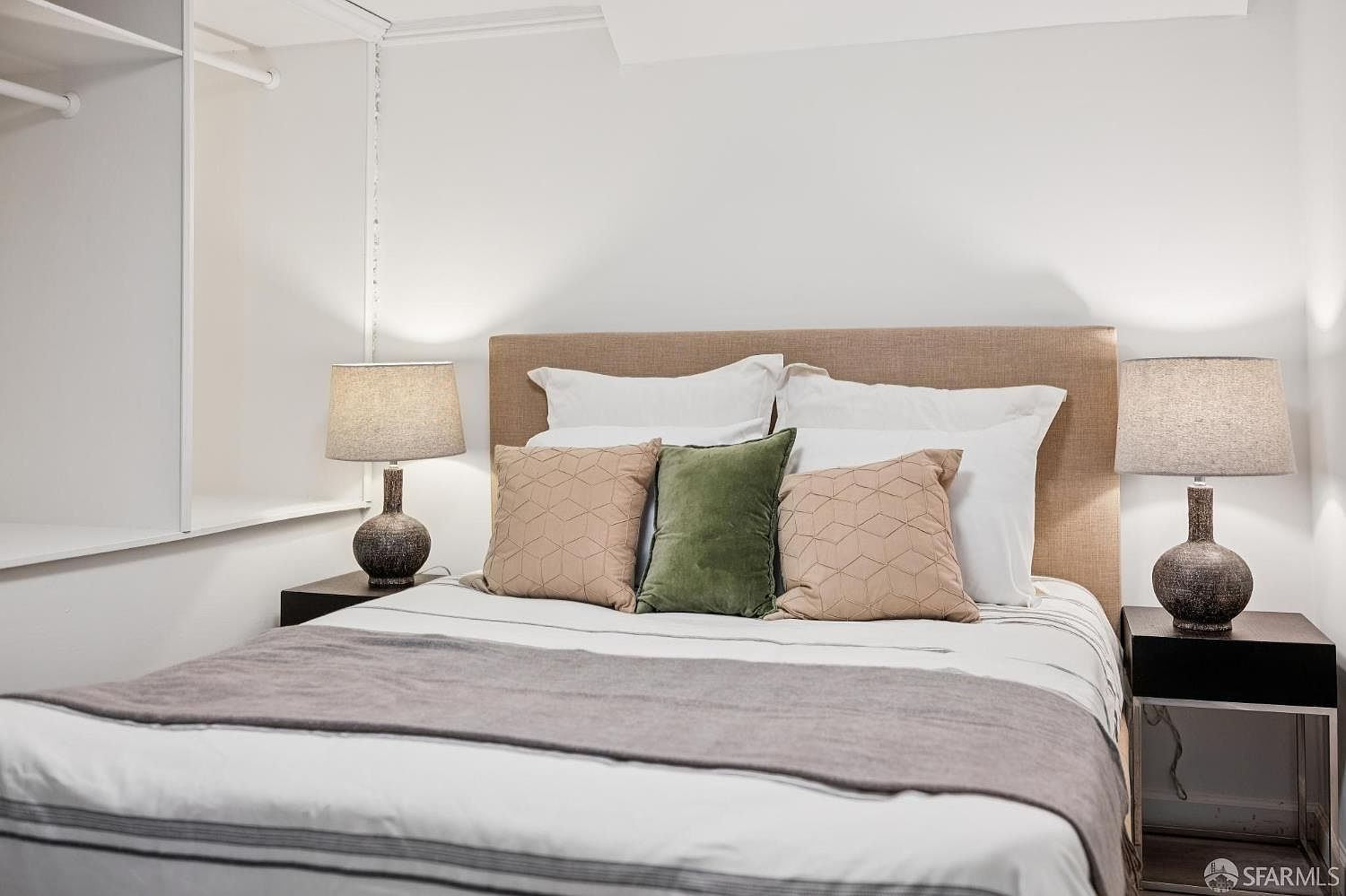 This cozy bedroom features a centered bed with a neutral-toned upholstered headboard, dressed in crisp white linens and a grey throw blanket. The bed is accented by three decorative pillows in shades of tan and olive green, flanked by two matching dark-toned bedside tables topped with textured, spherical table lamps. To the left, a built-in shelving unit with a hanging rod provides organized storage, creating a clean and functional sleeping space.