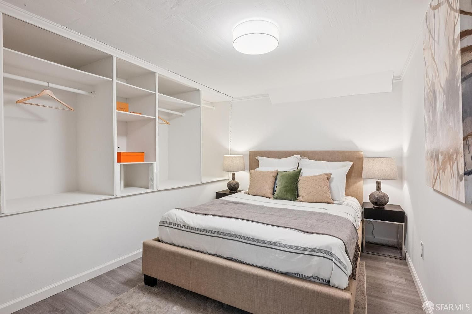 This cozy bedroom features a neutral color palette with a beige upholstered bed frame and matching headboard. To the left, a built-in white shelving unit provides open storage and hanging space, while two bedside tables with matching lamps flank the bed. The room is finished with light-toned wood-look flooring and a large piece of abstract wall art, creating a clean and inviting atmosphere.