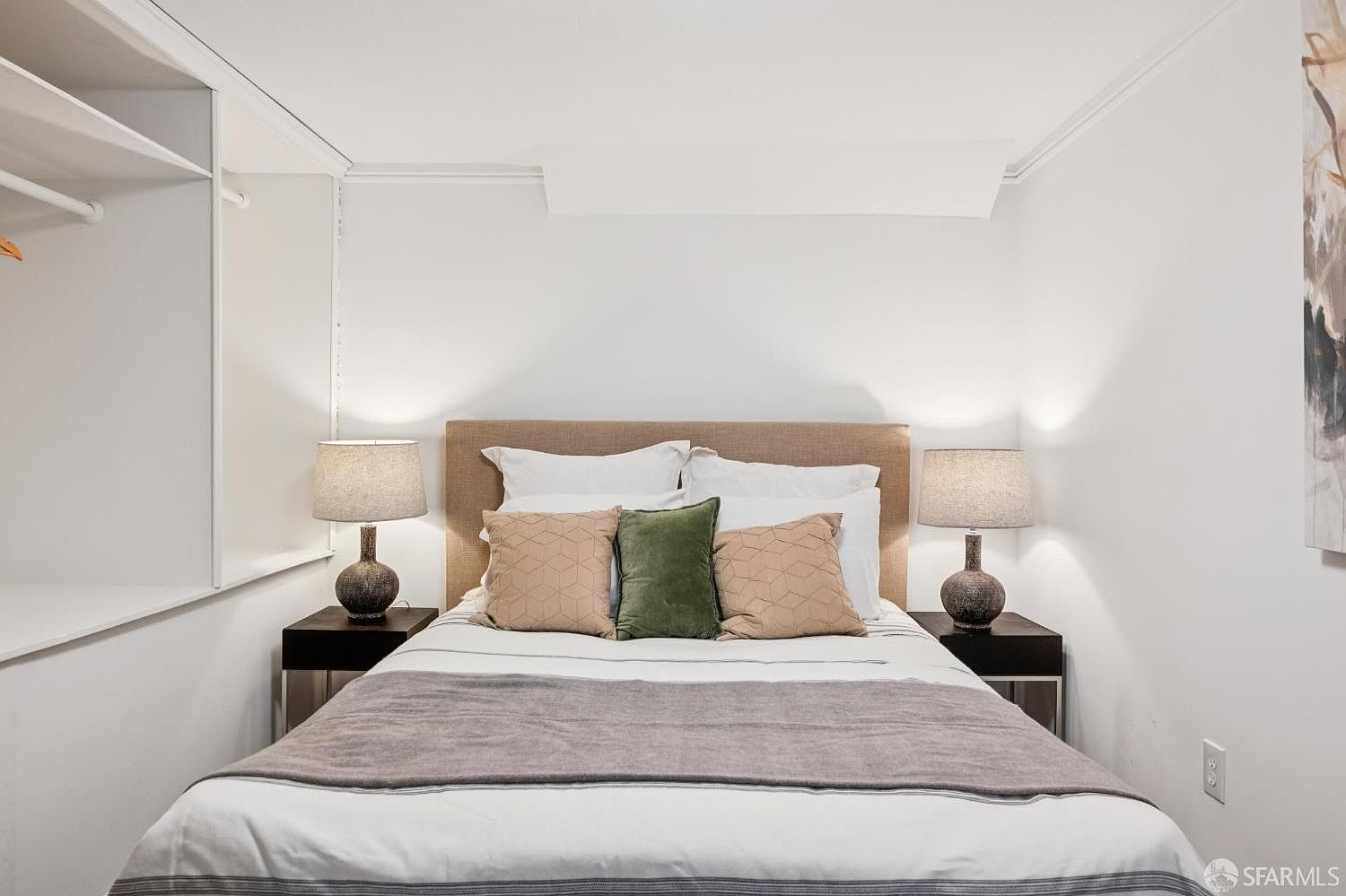 This cozy bedroom features a centered bed with a neutral-toned upholstered headboard, flanked by two matching dark wood nightstands and textured table lamps. The bed is dressed in crisp white linens, accented by a gray throw blanket and a trio of decorative pillows in tan and olive green. The room is framed by white walls, with a built-in closet area visible on the left and a piece of abstract wall art on the right, creating a clean and inviting atmosphere.