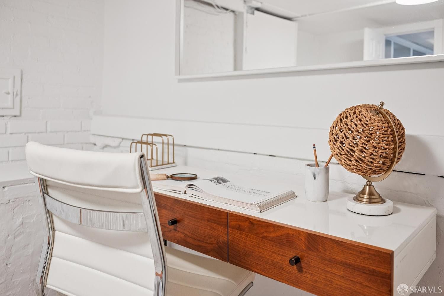 This bright and minimalist home office space features a sleek desk with a white top and two wood-toned drawers, paired with a modern white chair. The desk is accessorized with a decorative woven globe, a pencil holder, and an open book, set against a white painted brick wall. A large mirror hangs above the desk, reflecting the clean, contemporary aesthetic of the room.