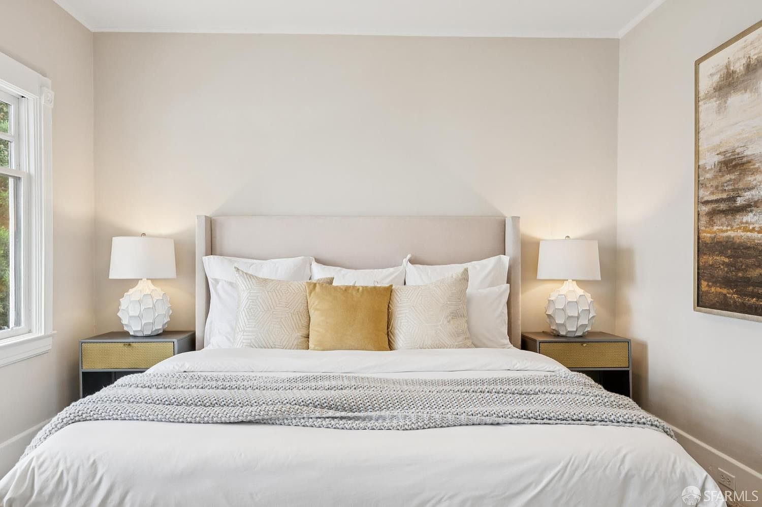 This serene primary bedroom features a neutral color palette with a plush, upholstered headboard as the central focal point. The bed is dressed in crisp white linens, accented by a textured gray throw blanket and a trio of decorative pillows in shades of gold and cream. Flanking the bed are two modern nightstands topped with geometric, white-textured table lamps, while a large piece of abstract wall art adds a sophisticated touch to the right side of the room.