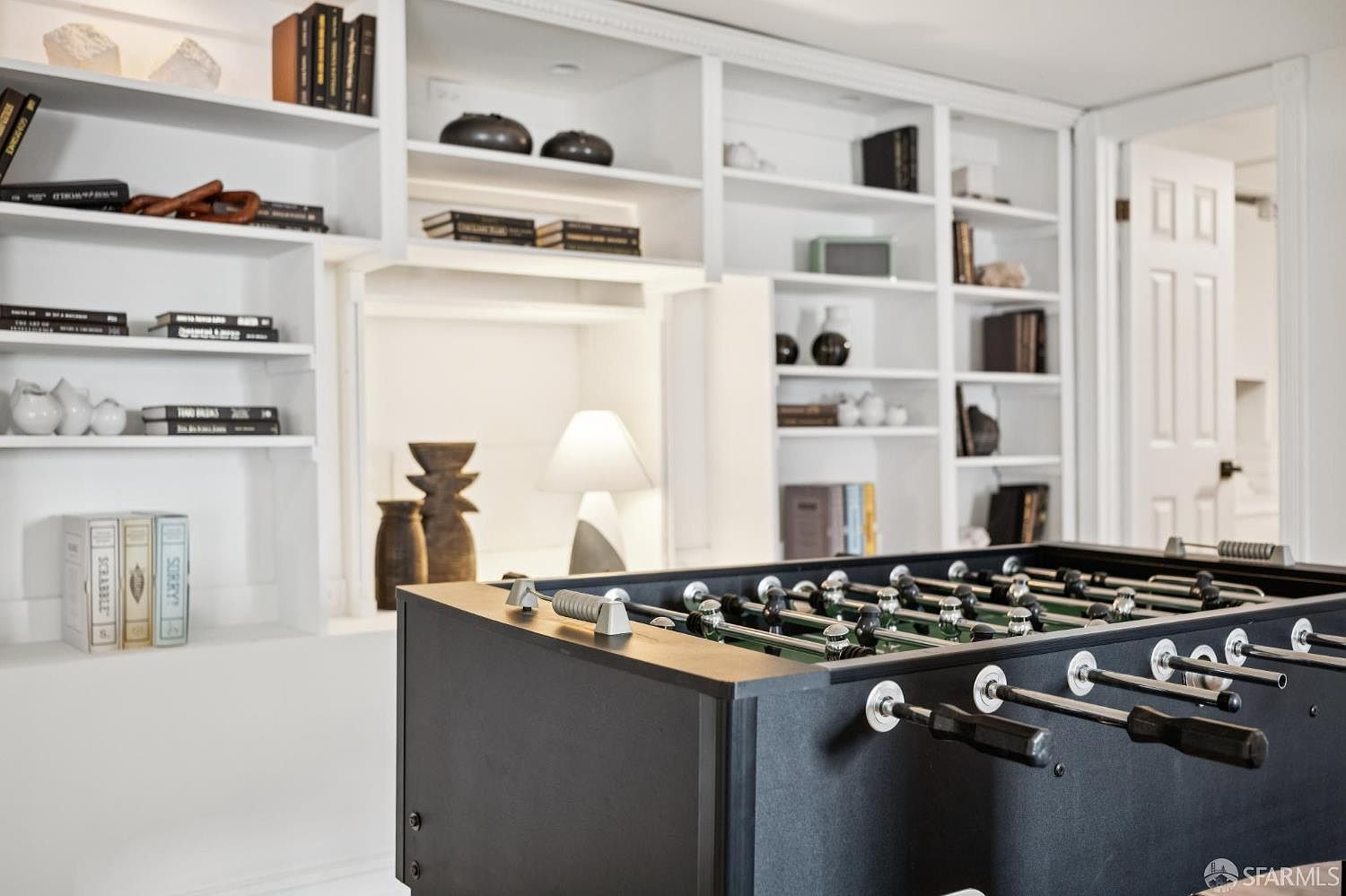 This interior space features a prominent black foosball table in the foreground, set against a backdrop of extensive white built-in shelving. The shelves are neatly organized with books, decorative objects, and a table lamp, creating a sophisticated and recreational atmosphere. The perspective captures the contrast between the dark game table and the bright, clean lines of the storage units, suggesting a versatile room for leisure or work.