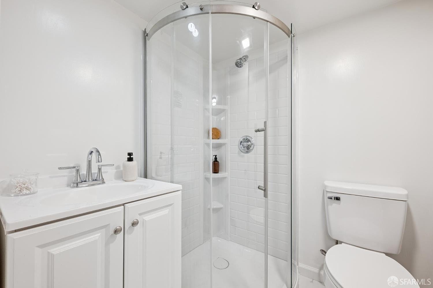 This clean, compact bathroom features a white vanity with a chrome faucet and a curved glass-enclosed shower with white subway tile walls. The space is bright and minimalist, utilizing a neutral color palette to maximize the sense of openness. A standard white toilet is positioned to the right, completing this functional and well-maintained bathroom layout.