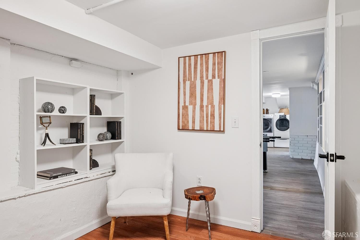This cozy, well-lit basement nook features a white armchair placed beside a built-in bookshelf and a unique, rustic wood-topped side table. A large, abstract geometric art piece hangs on the wall, while a doorway leads into an adjacent laundry area equipped with a washer and dryer. The space is finished with light wood flooring and crisp white walls, creating a clean and inviting atmosphere.