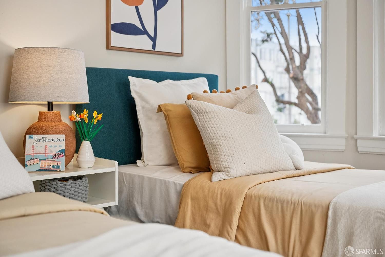 This cozy bedroom features a twin bed with a deep teal headboard, layered with mustard yellow and white bedding. A bedside table holds a textured lamp and a decorative San Francisco-themed book, while a small vase of artificial flowers adds a touch of color. The room is brightened by natural light streaming through a window that overlooks a tree outside.
