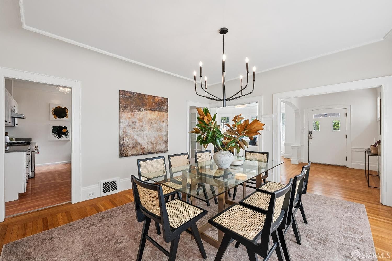 This elegant dining room features a glass-topped table surrounded by six black-framed chairs with woven cane seats, set atop a neutral-toned area rug. A modern, multi-armed chandelier hangs above a central vase with large green leaves, while a large abstract painting adds a focal point to the wall. The space is bright and airy, with hardwood floors and open transitions leading to the kitchen and entryway.