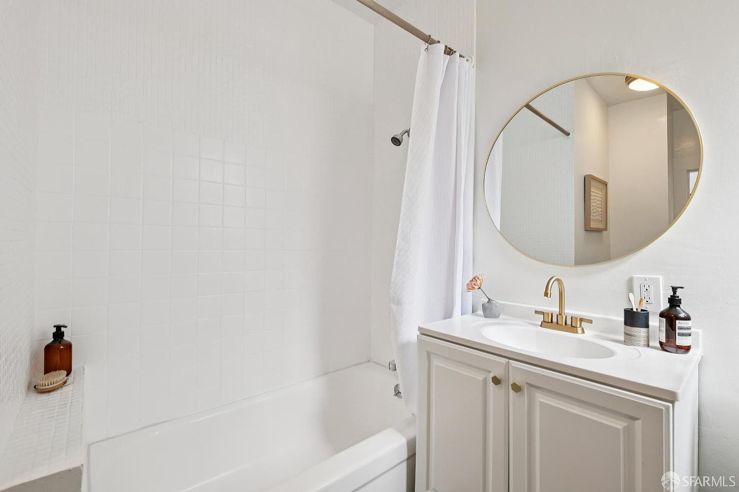 This bright and clean bathroom features a white vanity with a gold-toned faucet and a matching circular mirror. A white shower curtain hangs over a standard bathtub, which is tiled with white square tiles. The space is accented with a small vase of flowers and minimalist toiletries, creating a fresh and inviting atmosphere.