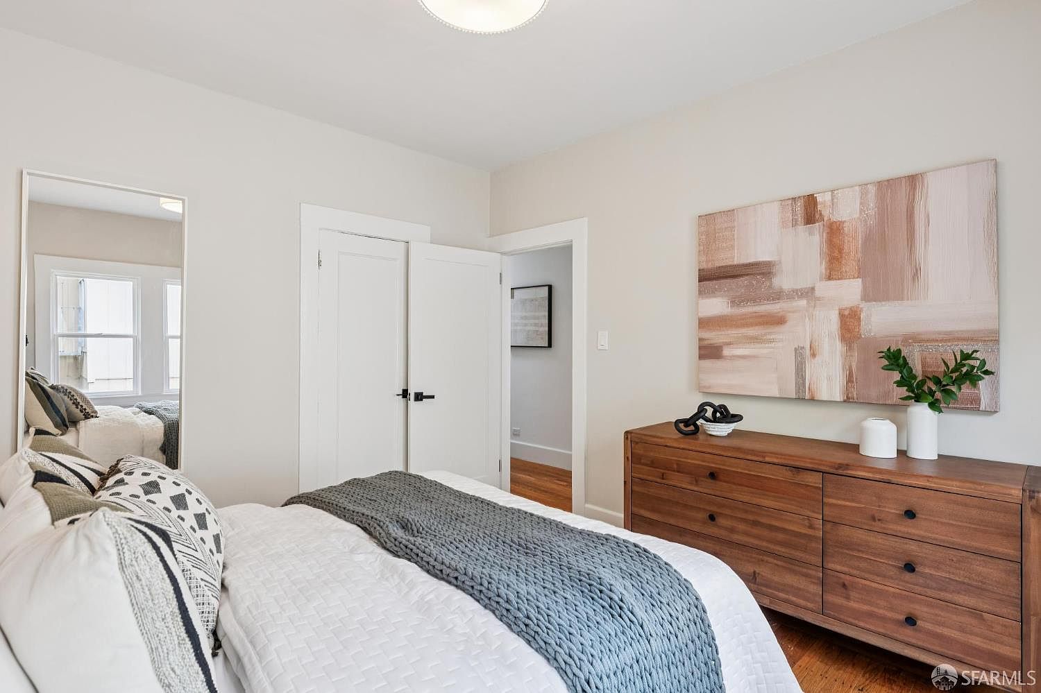 This bright and airy bedroom features a neutral color palette with a large wooden dresser topped with decorative vases and greenery. A textured abstract art piece hangs above the dresser, complementing the cozy bed dressed in white linens and a chunky grey knit throw. A tall floor mirror reflects the bed, while a doorway leads into an adjacent hallway, creating a sense of depth and flow in the space.