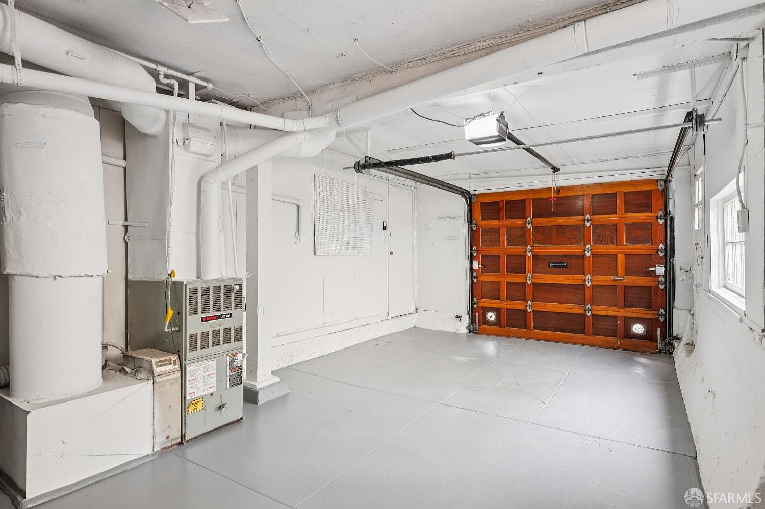 This interior view of a garage features a classic wooden sectional door with multiple panels and a central mail slot. The space is finished with painted white walls and a light gray floor, with exposed overhead pipes and a furnace unit positioned on the left side. A window on the right wall provides natural light, creating a functional and utilitarian storage or parking area.