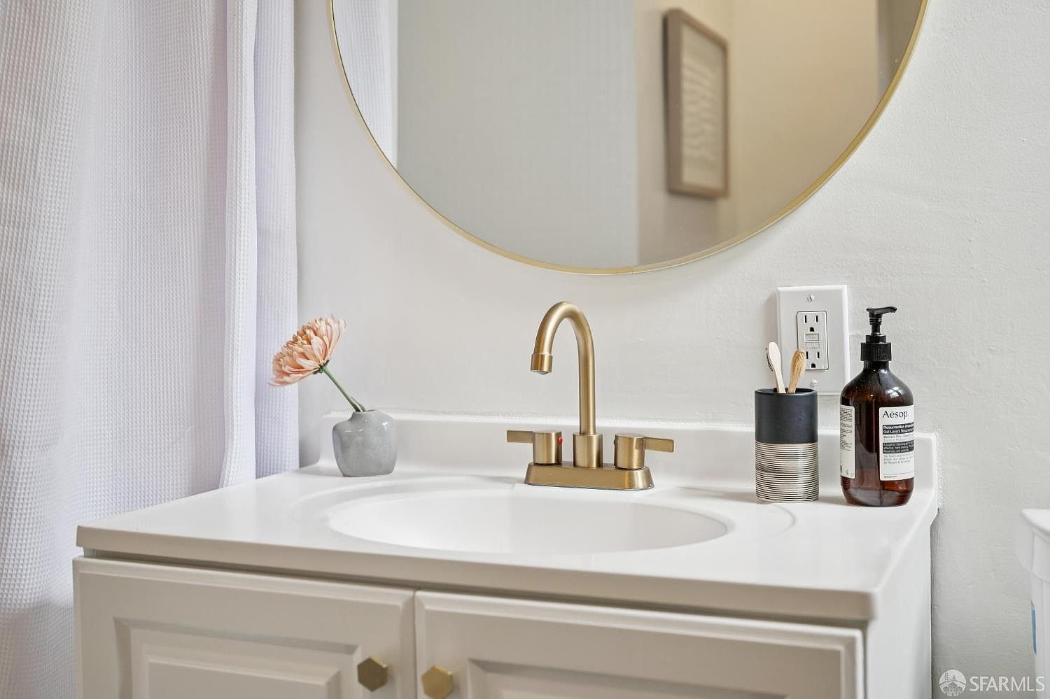 This bright and clean bathroom vanity features a white cabinet base with gold-toned hardware, topped with a white countertop and an integrated sink. A modern gold faucet complements the circular mirror mounted on the wall above, while a small vase with a flower, a toothbrush holder, and a bottle of Aesop soap add a touch of curated style. The space is well-lit and presents a contemporary, minimalist aesthetic.