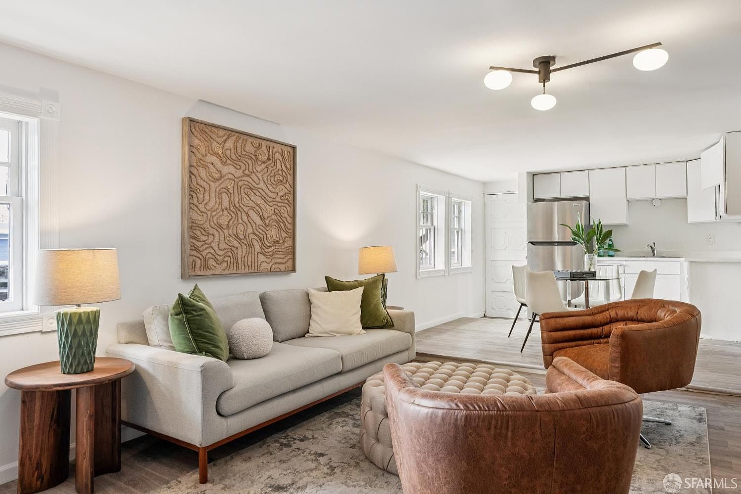 This open-concept living area features a light gray sofa accented with green and cream pillows, positioned on a textured area rug. Two brown leather swivel chairs and a tufted ottoman create a cozy seating arrangement, while a large, intricate wall art piece serves as a focal point above the sofa. The space flows seamlessly into a dining area and kitchen, characterized by neutral tones, modern light fixtures, and a bright, airy atmosphere.