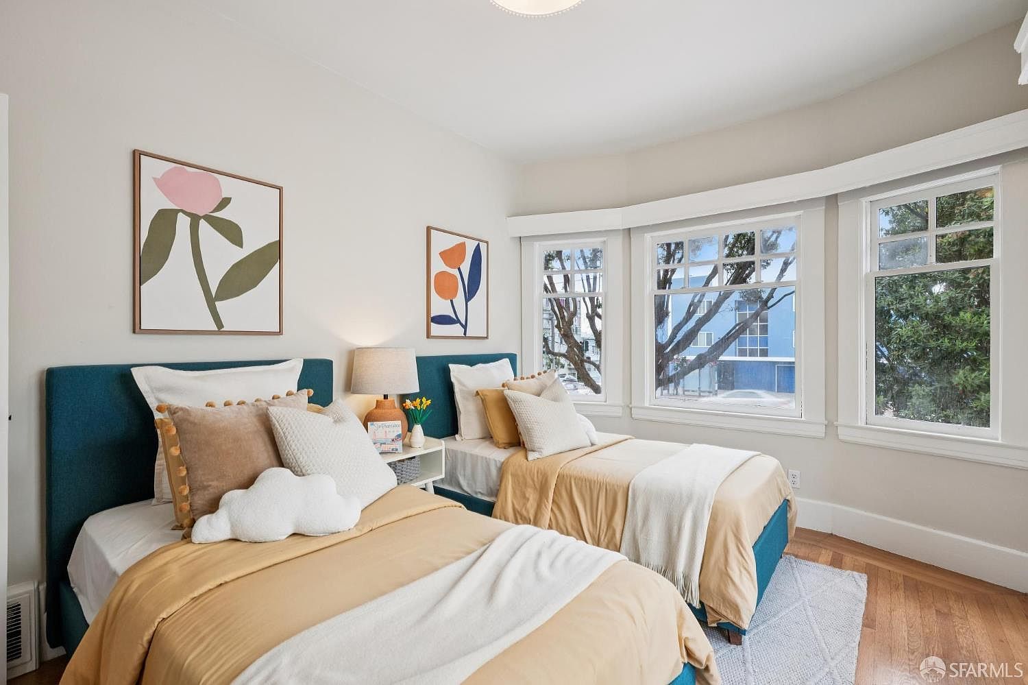 This bright and inviting guest bedroom features two twin beds with teal upholstered headboards and warm-toned bedding. The room is accented by minimalist floral wall art and a central nightstand with a lamp, while a large bay window allows for ample natural light and a view of the outdoors. The space is finished with light-colored walls and hardwood flooring, creating a cozy and well-lit atmosphere.
