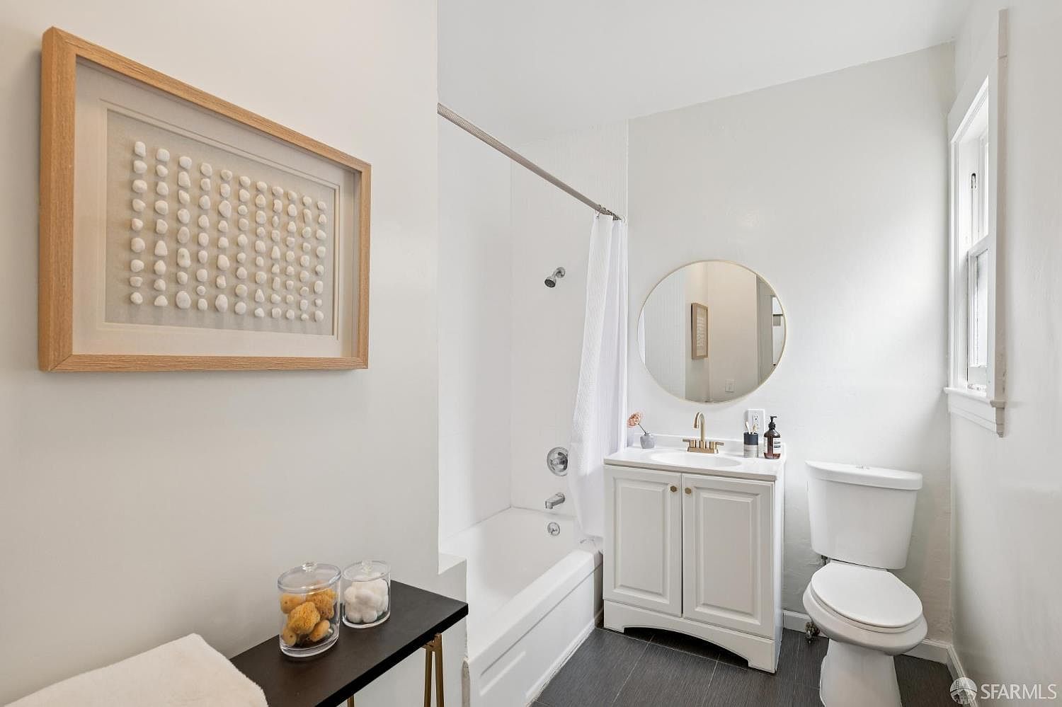 This bright, compact bathroom features a white vanity with a round mirror and a shower-tub combination with a white curtain. The space is finished with dark flooring and neutral walls, accented by a framed piece of textured wall art on the left. A small black console table in the foreground holds decorative jars, adding a touch of organization to the clean, functional layout.
