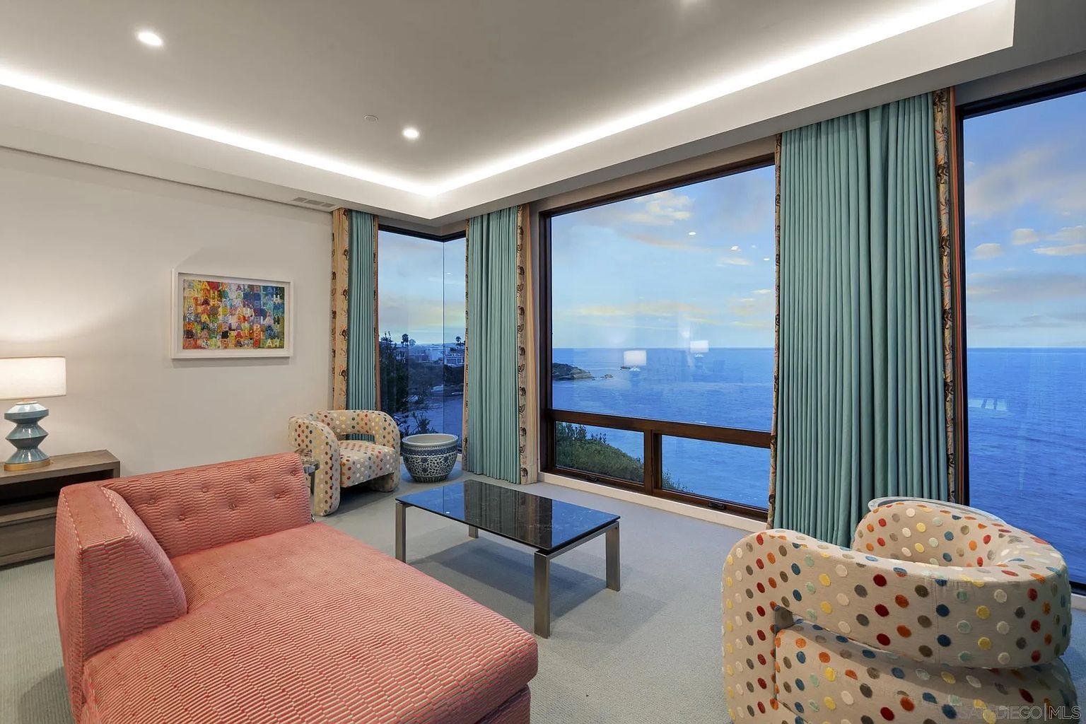 This bright living room features a contemporary aesthetic with a pink sofa and two patterned armchairs arranged around a glass-topped coffee table. The space is defined by expansive, large windows that offer a panoramic view of the ocean, framed by floor-to-ceiling teal curtains. Recessed lighting and a minimalist color palette create an airy, sophisticated atmosphere perfect for coastal living.