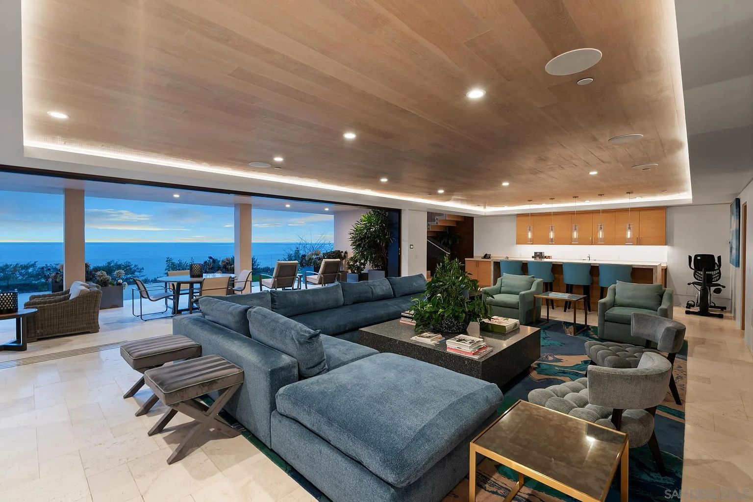 This expansive, open-concept living area features a large, plush blue sectional sofa centered around a substantial stone-topped coffee table. The space seamlessly transitions to an outdoor patio through wide, retractable glass walls that frame a panoramic ocean view. A bar area with light wood cabinetry and blue stools sits to the right, while the ceiling is finished with warm-toned wood planks and recessed lighting.