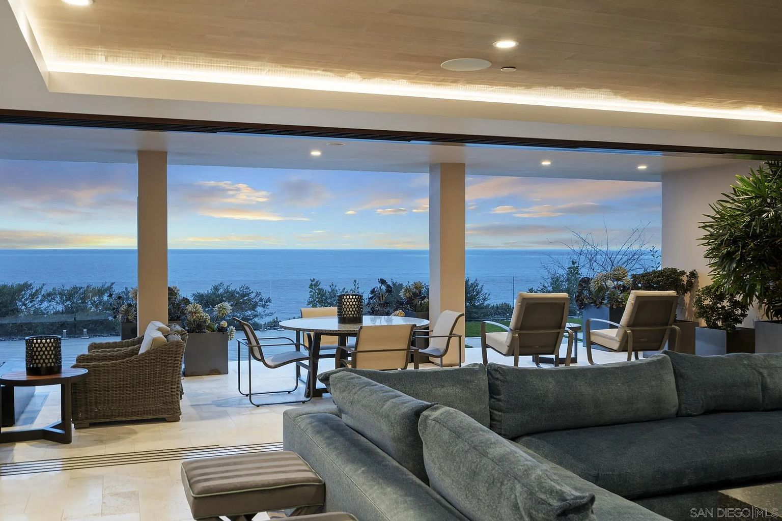 This expansive living area features a large, plush sectional sofa facing a seamless transition to an outdoor patio through wide-open glass walls. The space is defined by a neutral color palette, light wood-paneled ceilings with recessed lighting, and a direct, unobstructed view of the ocean. The patio area includes a dining set and comfortable lounge seating, creating a sophisticated indoor-outdoor living experience.