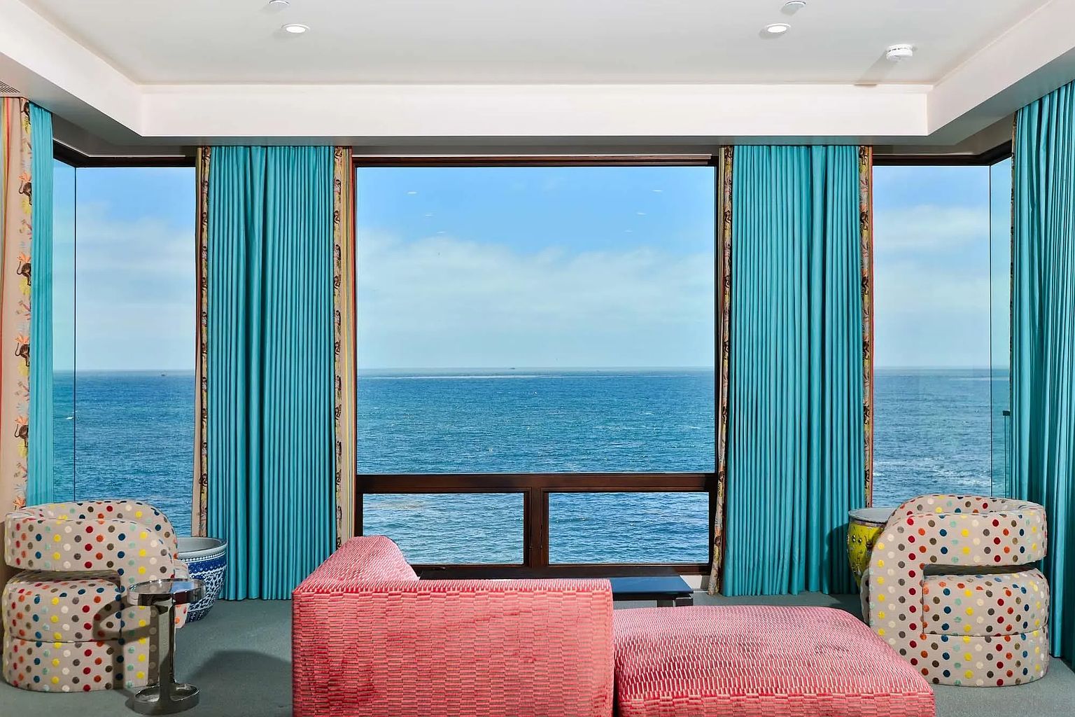 This vibrant living space features a striking panoramic view of the ocean framed by large windows and bold, pleated turquoise curtains. The room is furnished with two patterned, multi-colored armchairs and a textured, coral-toned sofa that anchors the seating area. The overall aesthetic is eclectic and colorful, emphasizing the dramatic coastal scenery as the primary focal point.