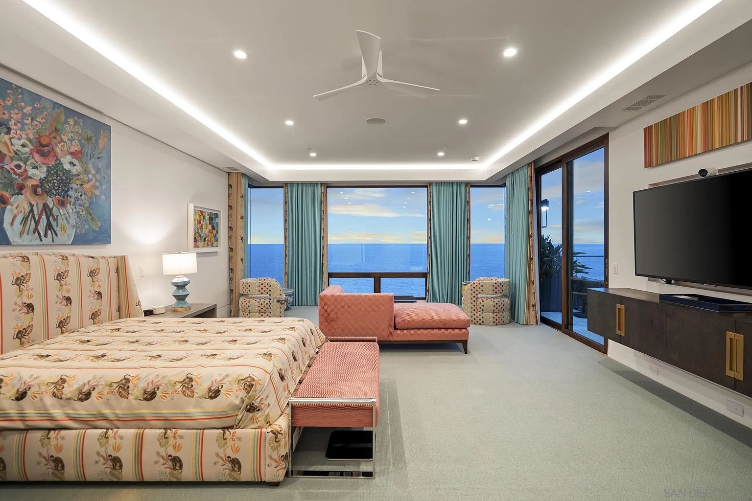 This spacious primary bedroom features a large bed with a patterned headboard and matching bedding, complemented by a pink upholstered bench at the foot. The room is anchored by a central seating area with a chaise lounge and two patterned armchairs, all positioned to take advantage of the expansive ocean views through large windows. A wall-mounted television sits above a dark wood media console, while recessed lighting and a modern ceiling fan provide ambient illumination throughout the space.