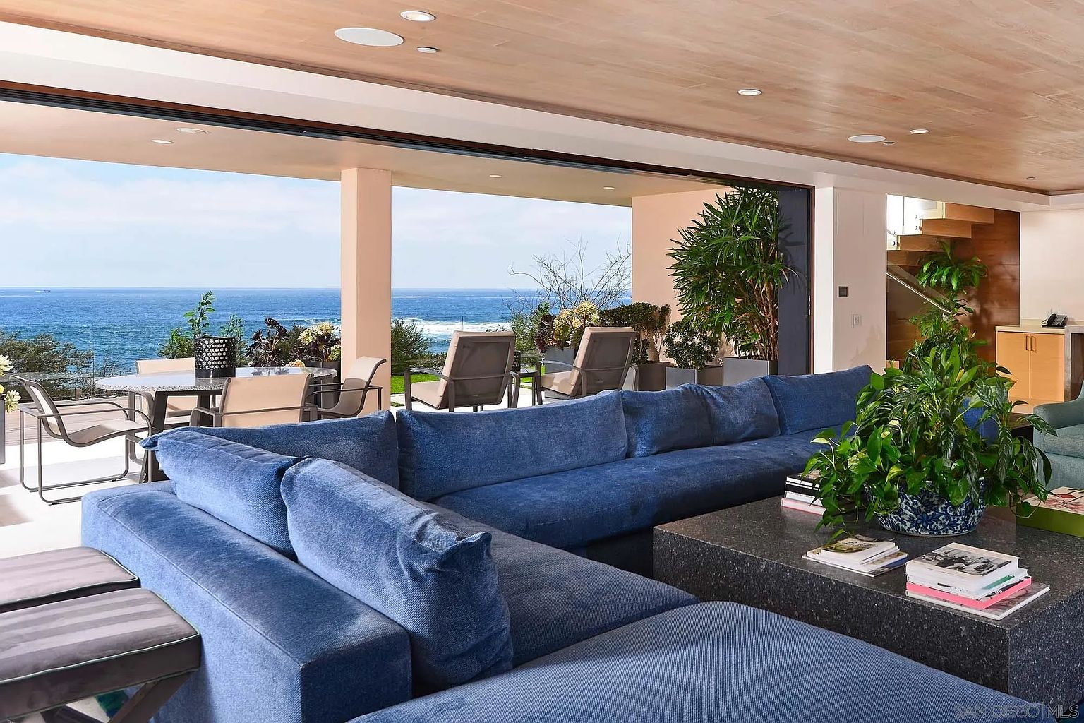 This expansive living room features a large, plush blue sectional sofa centered around a dark stone coffee table, creating a comfortable seating area. The space seamlessly transitions to an outdoor patio through wide, open glass walls that frame a panoramic view of the ocean. The interior is characterized by a light wood ceiling with recessed lighting, offering a bright and airy atmosphere that emphasizes the connection between indoor and outdoor living.