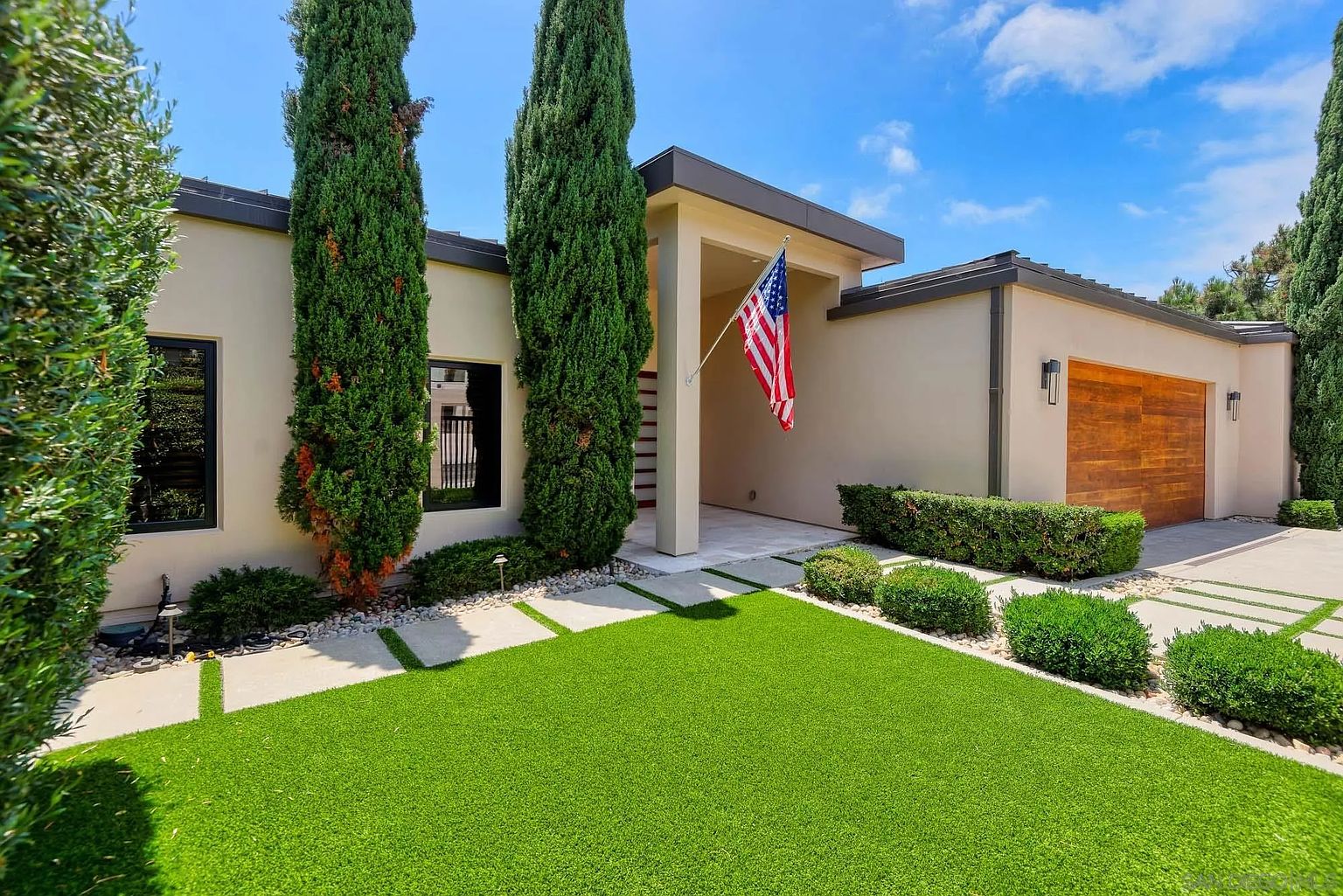 This modern single-story home features a clean, minimalist facade with smooth light-colored stucco walls and a prominent wooden garage door. The front entryway is sheltered by a deep overhang, accented by a proudly displayed American flag, while tall, slender cypress trees frame the entrance. A manicured artificial lawn with geometric concrete pavers leads to the front door, creating a crisp and inviting curb appeal.