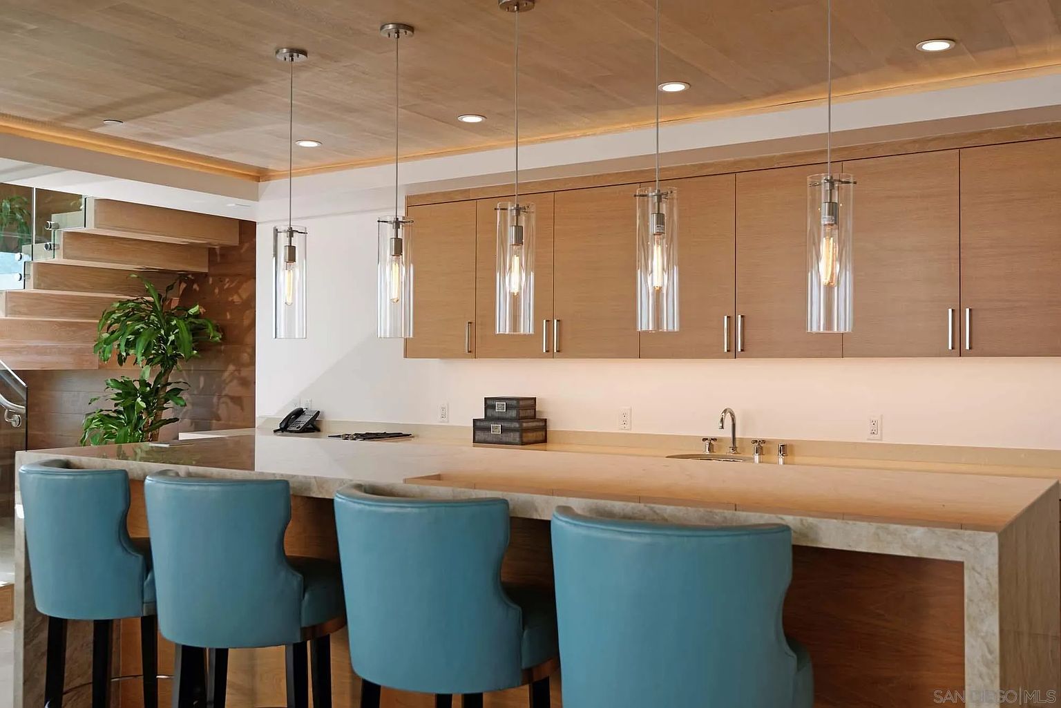 This modern kitchen features a spacious island with light-toned stone countertops and four vibrant blue bar stools, creating a welcoming breakfast bar area. Above the island, five sleek, cylindrical glass pendant lights hang from a warm-toned wood-paneled ceiling, while light wood cabinetry provides ample storage along the back wall. The perspective captures the clean lines of the kitchen workspace, with a glimpse of a wooden staircase and a potted plant in the background, emphasizing a contemporary and airy aesthetic.