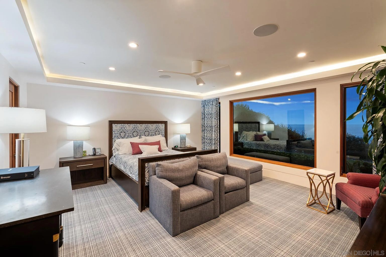 This spacious primary bedroom features a large bed with a patterned headboard, flanked by two nightstands with matching lamps. In the foreground, two gray upholstered armchairs are positioned facing the bed, while a large window provides a scenic view of the outdoors. The room is finished with neutral walls, a patterned carpet, and recessed lighting, creating a calm and sophisticated atmosphere.