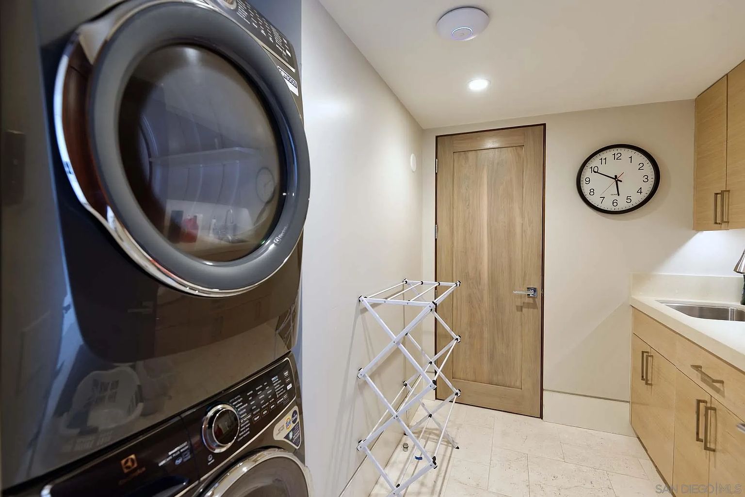 This functional laundry room features a stacked washer and dryer unit on the left and a light wood cabinet vanity with a sink on the right. A white folding drying rack stands in the center of the room, which is finished with light-colored tile flooring and a wall-mounted analog clock. The space is illuminated by recessed lighting and includes a wooden door leading to another area of the home.