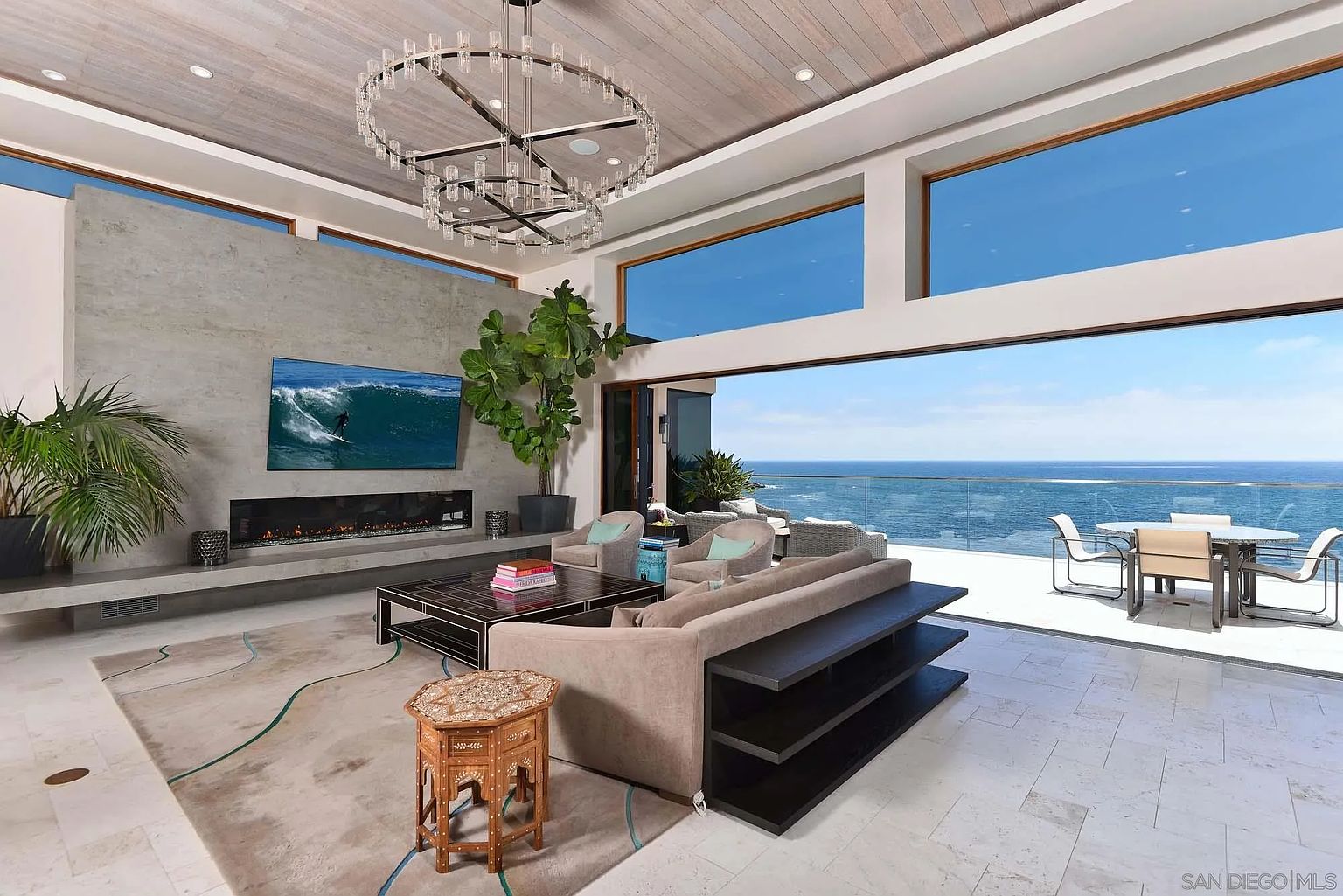 This expansive, modern living room features a neutral color palette and a seamless transition to an outdoor terrace overlooking the ocean. The space is anchored by a large, light-toned sofa, a dark coffee table, and a tall, stone-clad chimney breast housing a linear fireplace. A striking, multi-tiered circular chandelier hangs from the wood-paneled ceiling, while large glass panels provide an unobstructed, panoramic view of the sea.