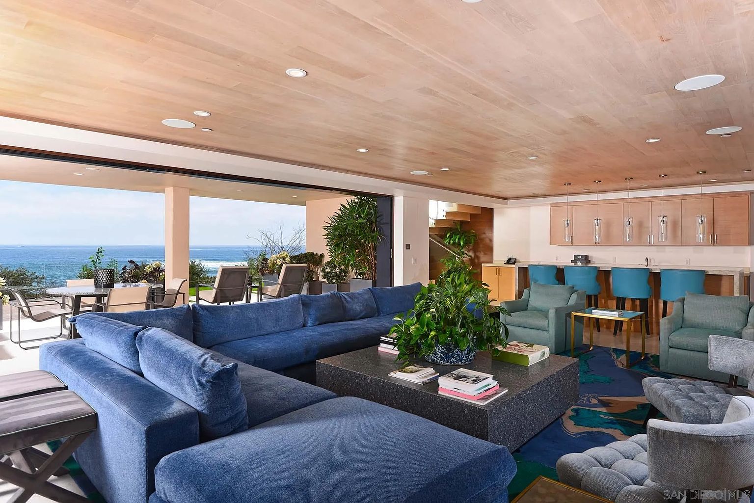 This expansive, open-concept living area features a large, plush blue sectional sofa centered around a substantial dark stone coffee table. The room seamlessly transitions to an outdoor patio through wide, retractable glass walls that frame a panoramic ocean view. A bar area with blue stools sits to the right, while the ceiling is finished in light wood paneling with recessed lighting, creating a sophisticated and airy coastal atmosphere.