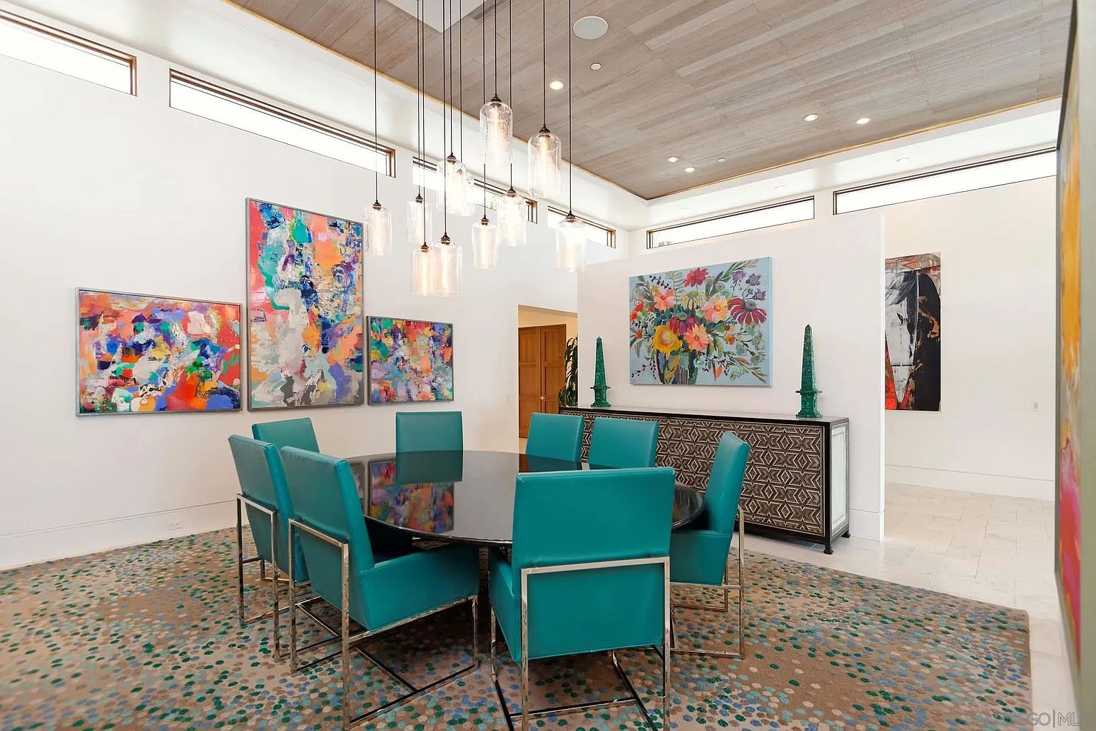This vibrant dining room features a dark, circular table surrounded by six bright teal chairs with metallic frames, set against a backdrop of colorful abstract artwork. A cluster of glass pendant lights hangs above the table, while a patterned sideboard sits against the wall, topped with two tall green decorative obelisks. The space is defined by a light wood-paneled ceiling and a large, speckled area rug, creating a bold and artistic atmosphere.