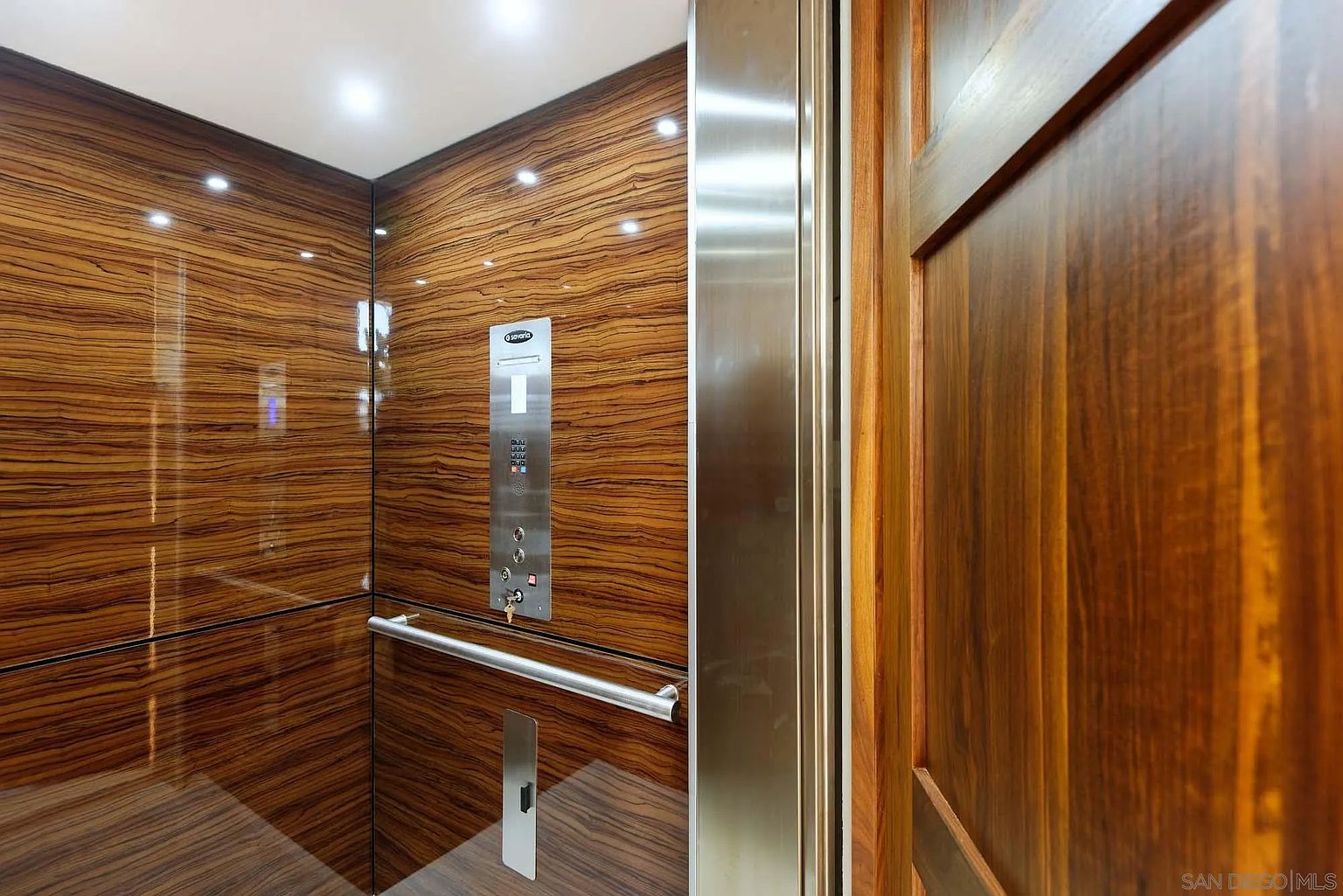 This image shows the interior of a residential elevator featuring high-gloss, wood-grain paneling on the walls. A central stainless steel control panel includes buttons and a key switch, complemented by a horizontal metal handrail and a matching metal threshold. The perspective is from inside the elevator looking toward the open doorway, highlighting the sleek, modern design and premium finishes.