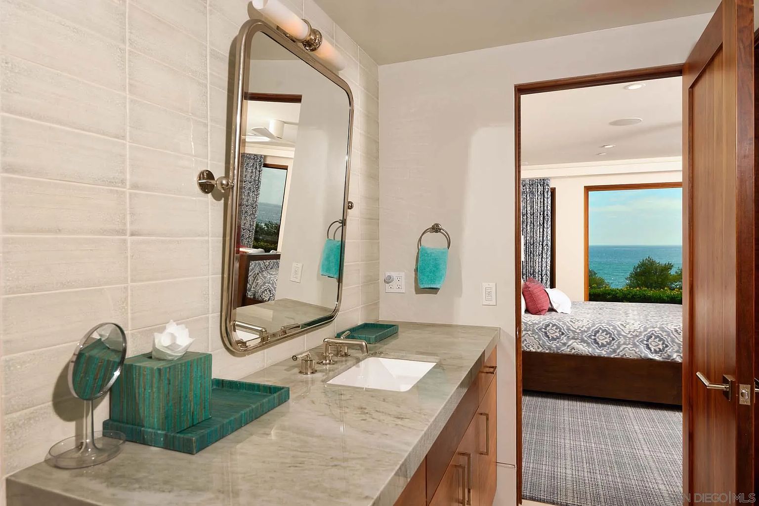This bathroom features a stone-topped vanity with a rectangular undermount sink and a large, framed mirror mounted on a textured, light-colored tiled wall. A doorway provides a direct line of sight into an adjacent bedroom, which boasts a view of the ocean through a large window. The space is accented with teal-colored accessories, creating a serene and cohesive aesthetic.
