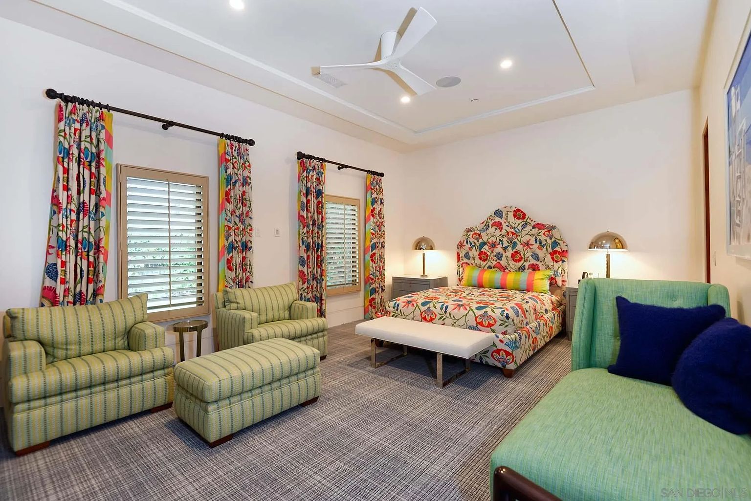 This vibrant bedroom features a bold floral-patterned bed frame and matching curtains that add a playful, eclectic touch to the space. The room is furnished with two striped armchairs, a matching ottoman, and a green chaise lounge, all set against a neutral backdrop with patterned carpeting. A white ceiling fan hangs above, and two bedside lamps provide warm lighting, creating a cozy and inviting atmosphere.