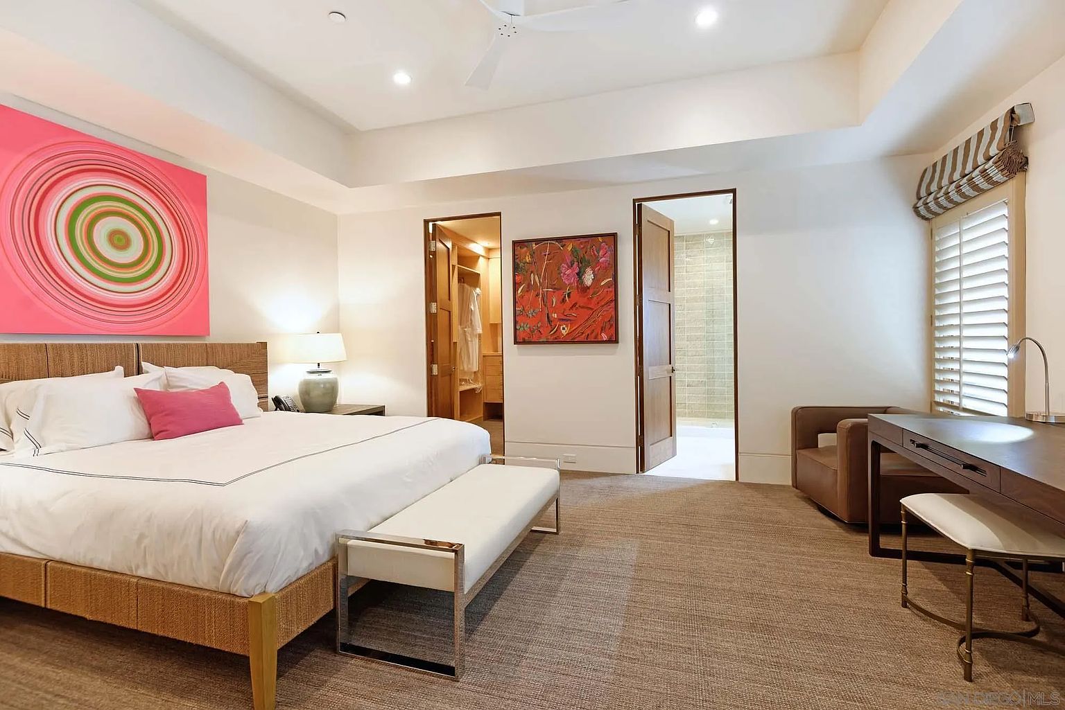 This spacious primary bedroom features a neutral color palette accented by vibrant, abstract artwork and a bold pink throw pillow. The room includes a large bed with a textured headboard, a sleek bench at the foot, and a dedicated desk area near the window with plantation shutters. Two doorways lead to a walk-in closet and a bathroom, creating a functional and well-appointed suite.