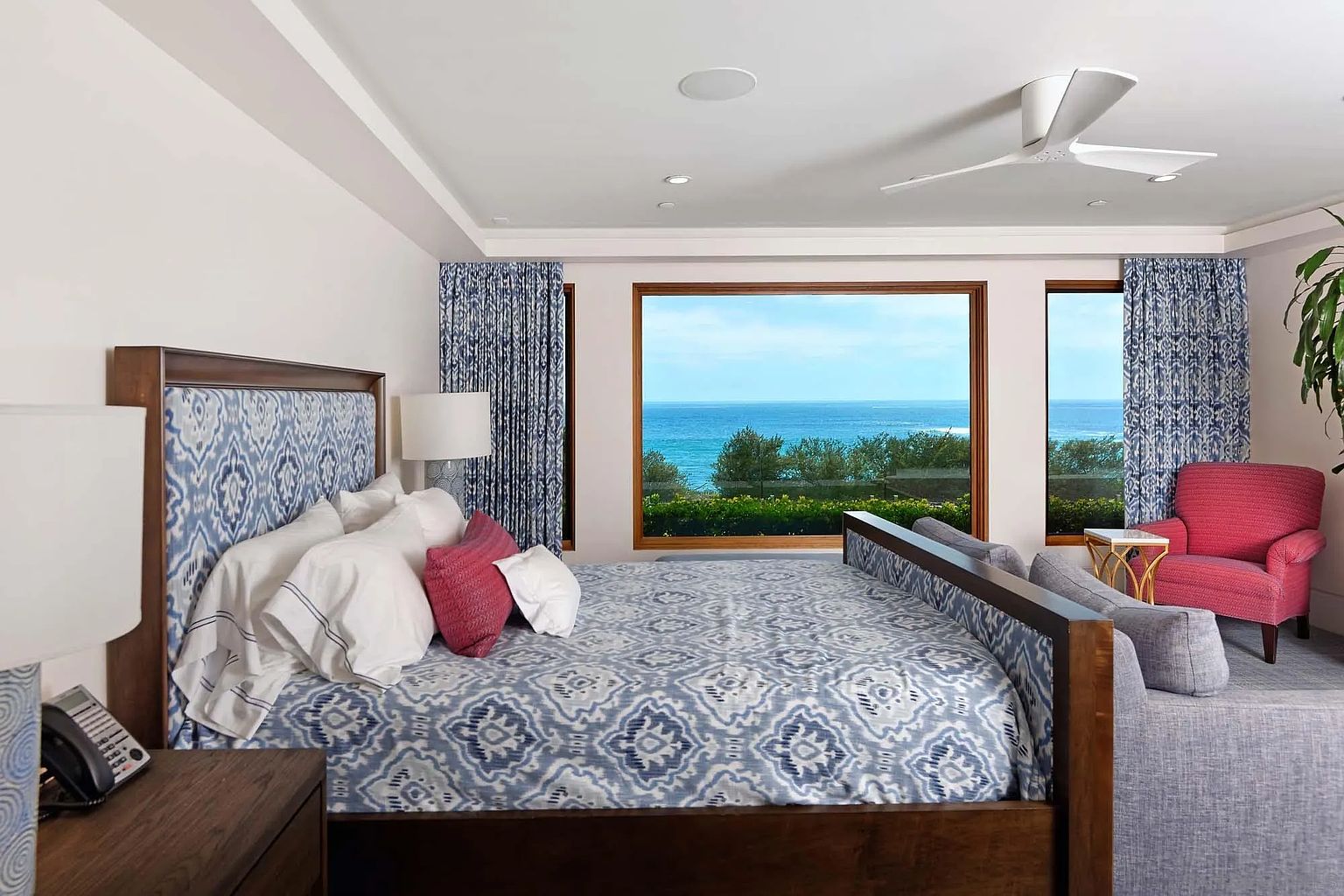 This primary bedroom features a large wooden bed frame with a patterned blue and white headboard, complemented by matching bedding and accent pillows. A large window provides a panoramic view of the ocean, while a cozy sitting area with a pink armchair and a gray sofa is positioned nearby. The room is illuminated by recessed lighting and a modern ceiling fan, creating a serene and luxurious atmosphere.