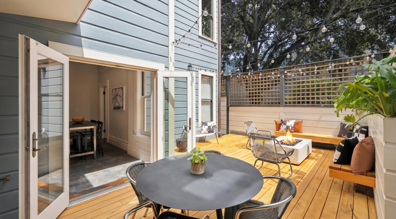 This inviting outdoor deck features a modern, multi-functional living space with a round dining table in the foreground and a cozy lounge area with a fire pit in the background. The deck is framed by light-colored wooden siding and a decorative lattice fence, accented by string lights that create a warm, ambient atmosphere. The perspective is a wide-angle shot from the deck looking toward the house, highlighting the seamless indoor-outdoor transition through open French doors.