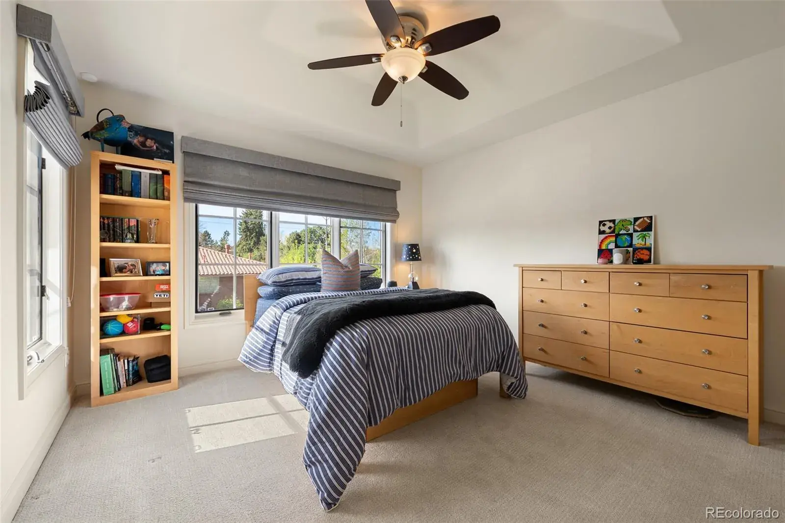 This is a well-lit bedroom featuring a bed with striped bedding and a dark throw, a wooden dresser with artwork above, and a bookshelf filled with various items. Natural light streams in through a large window with gray roman shades, and a ceiling fan adds to the room's comfort. The overall impression is a cozy and inviting space.