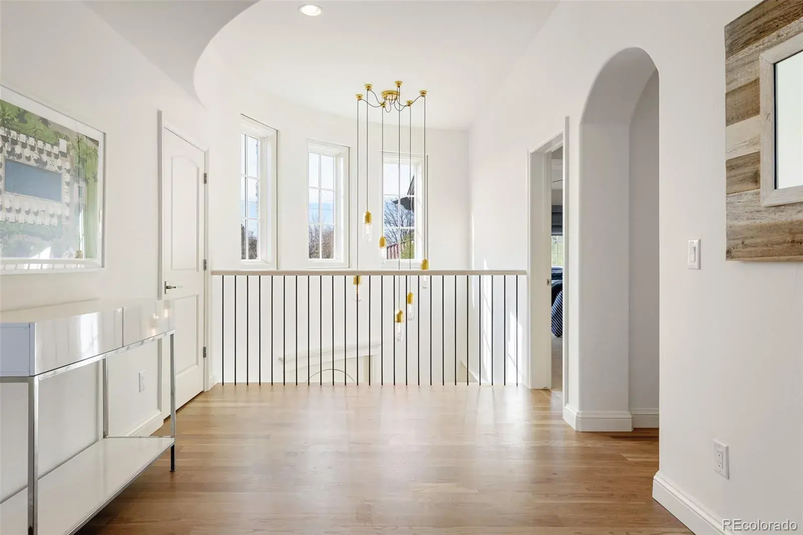 This interior shot showcases a bright and airy hallway with hardwood floors and white walls. A modern chandelier hangs above the staircase, which is lined with a white railing featuring black vertical bars. The space is illuminated by natural light streaming through several windows, creating a welcoming and elegant atmosphere.
