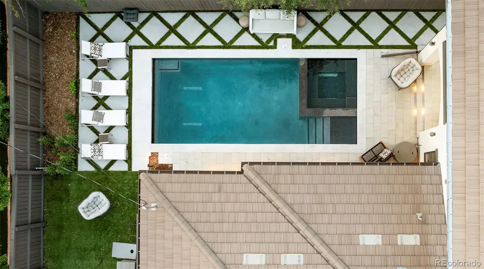 This aerial view showcases a luxurious backyard featuring a rectangular swimming pool with a spa, surrounded by a patio with lounge chairs and a seating area. The landscaping includes geometric stone pavers with grass accents, and the property is enclosed by a wooden fence. The roof of the house is visible, providing context to the outdoor space.