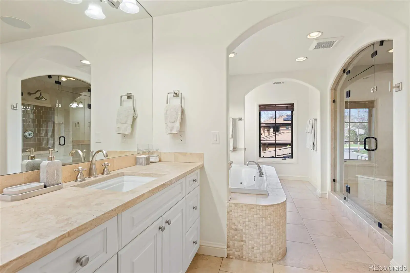 This is a well-lit primary bathroom featuring a vanity with a light-colored countertop and white cabinets. The bathroom includes a bathtub with mosaic tile accents and a glass-enclosed shower. The overall design is elegant and spacious, with neutral tones creating a relaxing atmosphere.