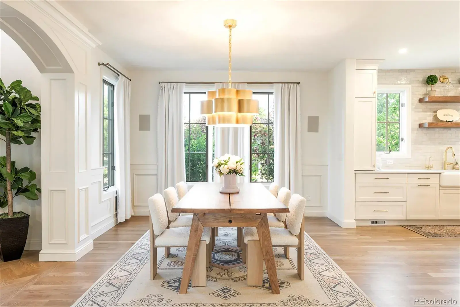 This is an interior shot of a dining room featuring a long wooden table surrounded by six chairs with white, textured upholstery. A modern, multi-tiered gold chandelier hangs above the table, which is adorned with a floral centerpiece. Large windows with white curtains provide natural light, and an area rug adds texture to the hardwood floors, creating a bright and inviting space.