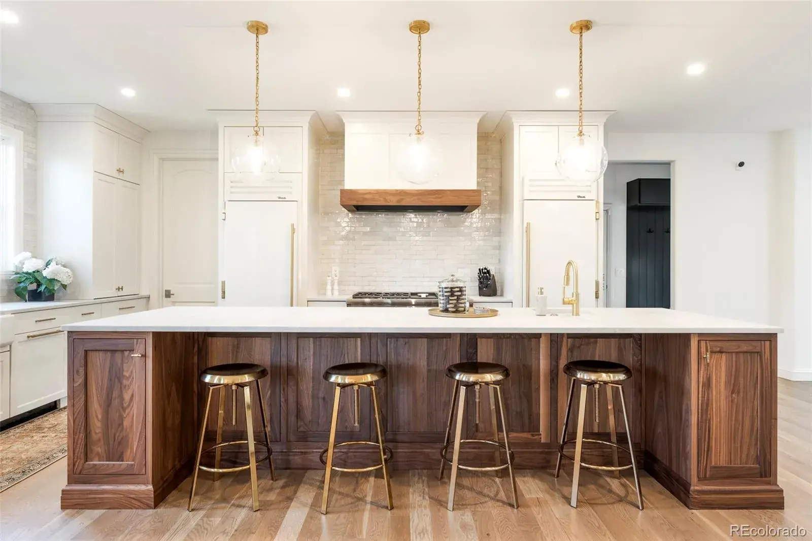 This is a well-lit kitchen featuring a large island with a white countertop and dark wood cabinetry. Four gold-colored bar stools are positioned along the island. The kitchen also includes white cabinets, stainless steel appliances, and pendant lighting, creating a modern and inviting space.