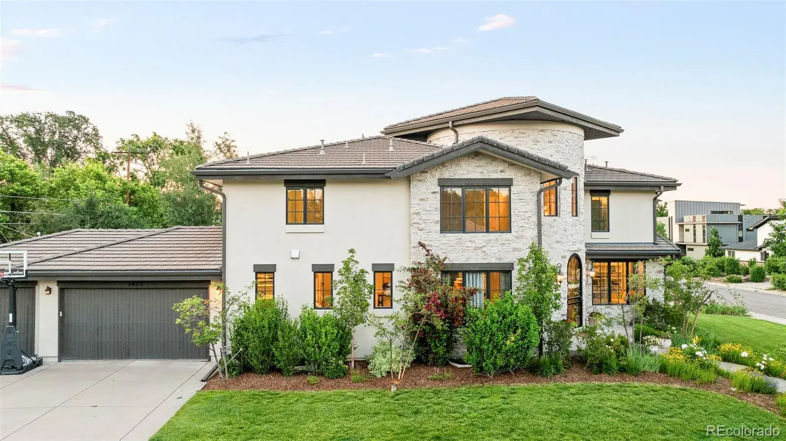 This is a front exterior view of a two-story house with a modern design. The house features a combination of stucco and stone facade, a tiled roof, and dark-framed windows. The well-manicured lawn and landscaping add to the property's curb appeal, and a basketball hoop is visible next to the garage.