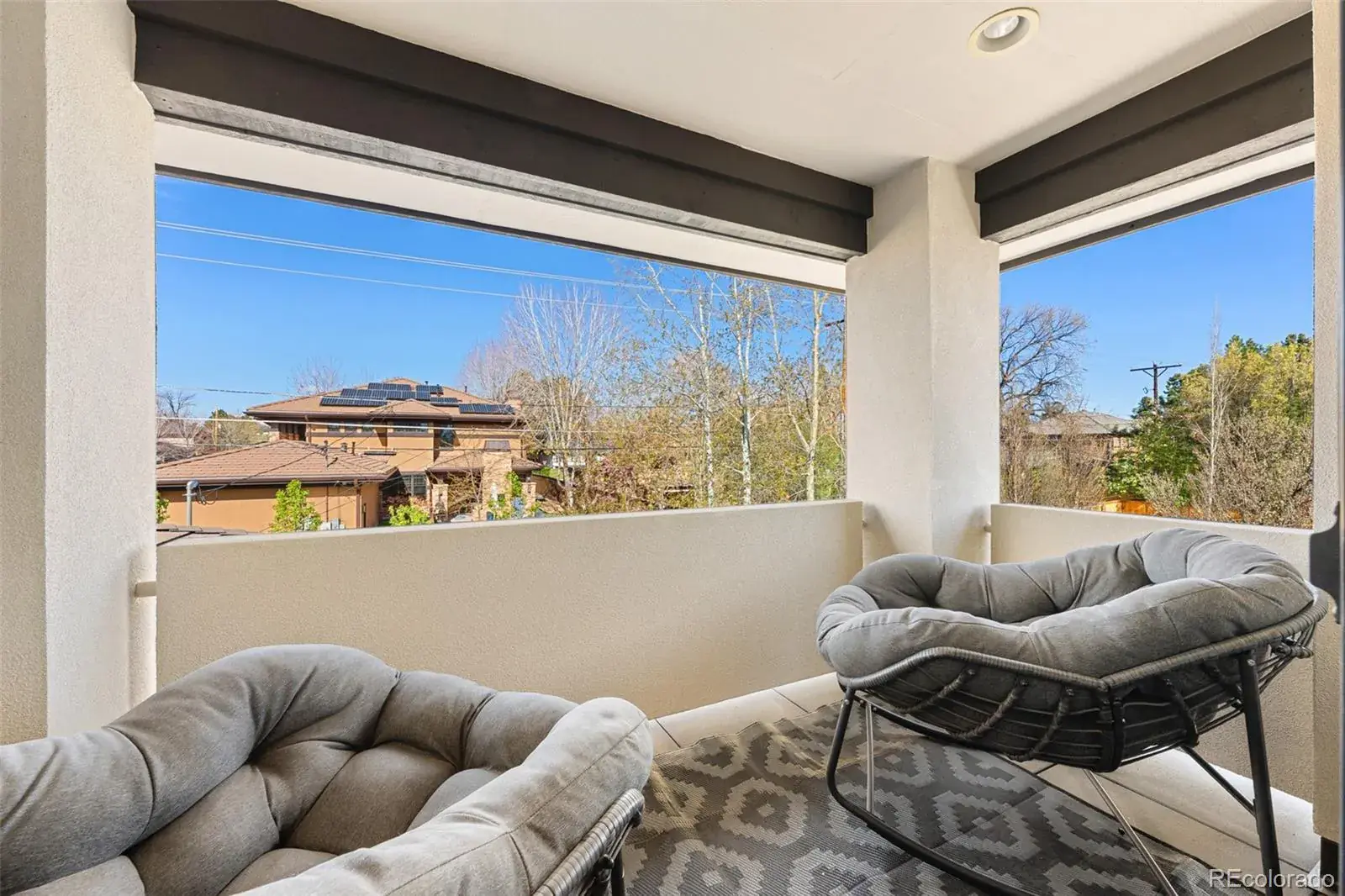 This image showcases a cozy balcony featuring two comfortable, oversized gray chairs arranged on a patterned gray rug. The balcony offers a view of neighboring houses and trees under a clear blue sky, framed by the balcony's structure with dark brown beams and white pillars. The space appears inviting and perfect for relaxation.