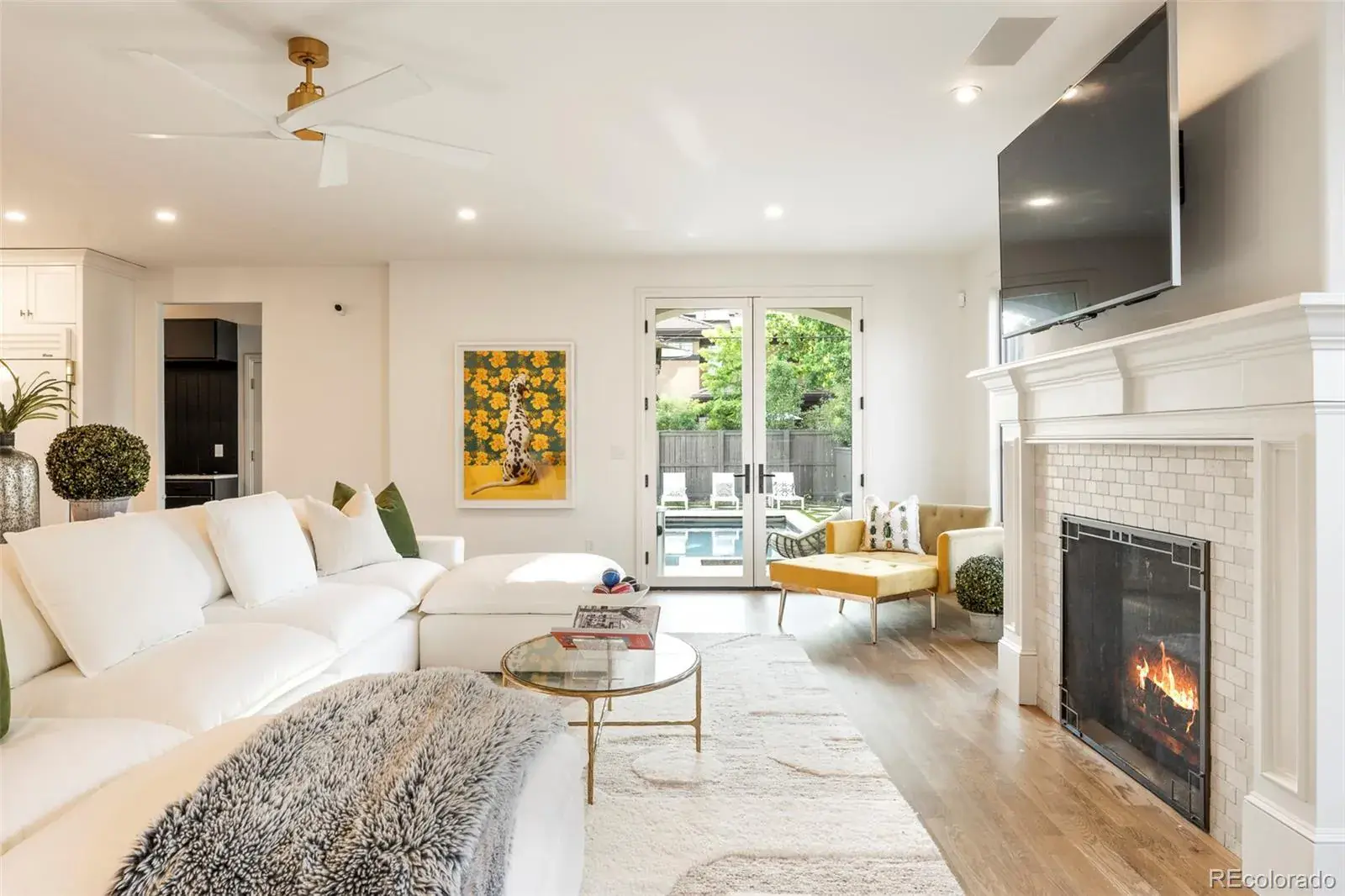 This is a bright and airy living room featuring a large white sectional sofa, a modern fireplace with a mounted television, and sliding glass doors leading to an outdoor pool area. The room is decorated in a contemporary style with hardwood floors, a round glass coffee table, and a colorful art piece, creating a welcoming and luxurious atmosphere.