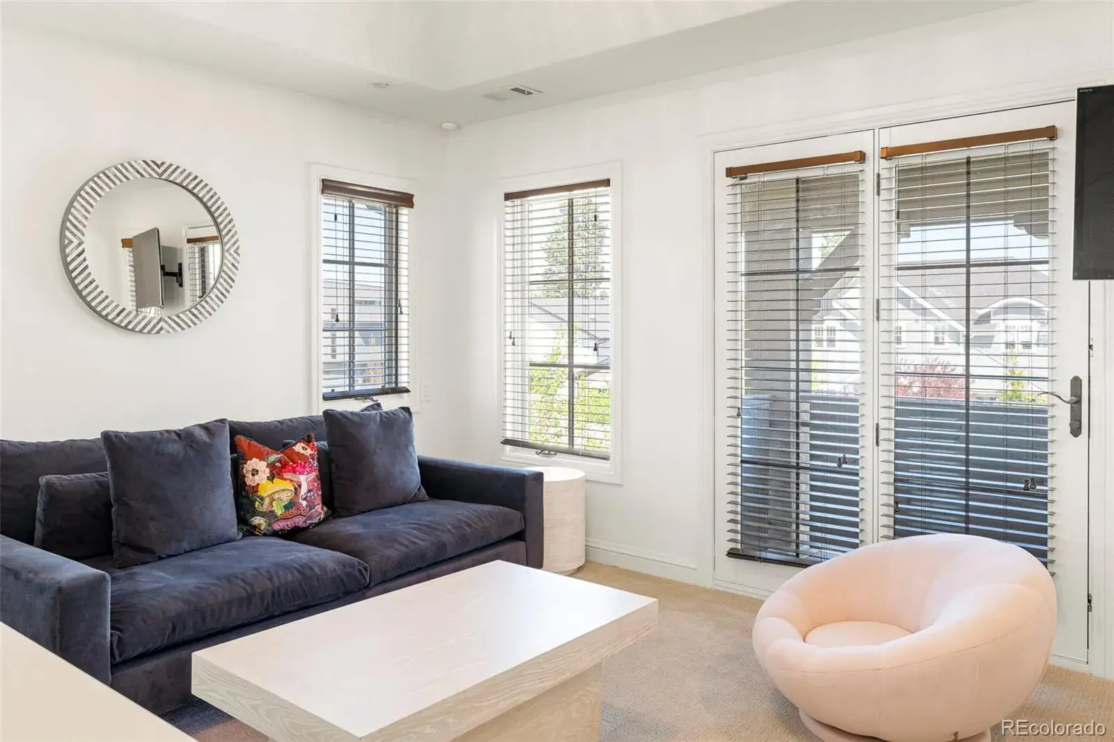 This is an interior shot of a living room featuring a dark blue sofa with decorative pillows, a light-colored coffee table, and a round pink chair. The room has multiple windows with blinds, allowing natural light to filter in, and a round mirror hangs on the wall, adding a decorative touch. The overall impression is a cozy and stylish living space.