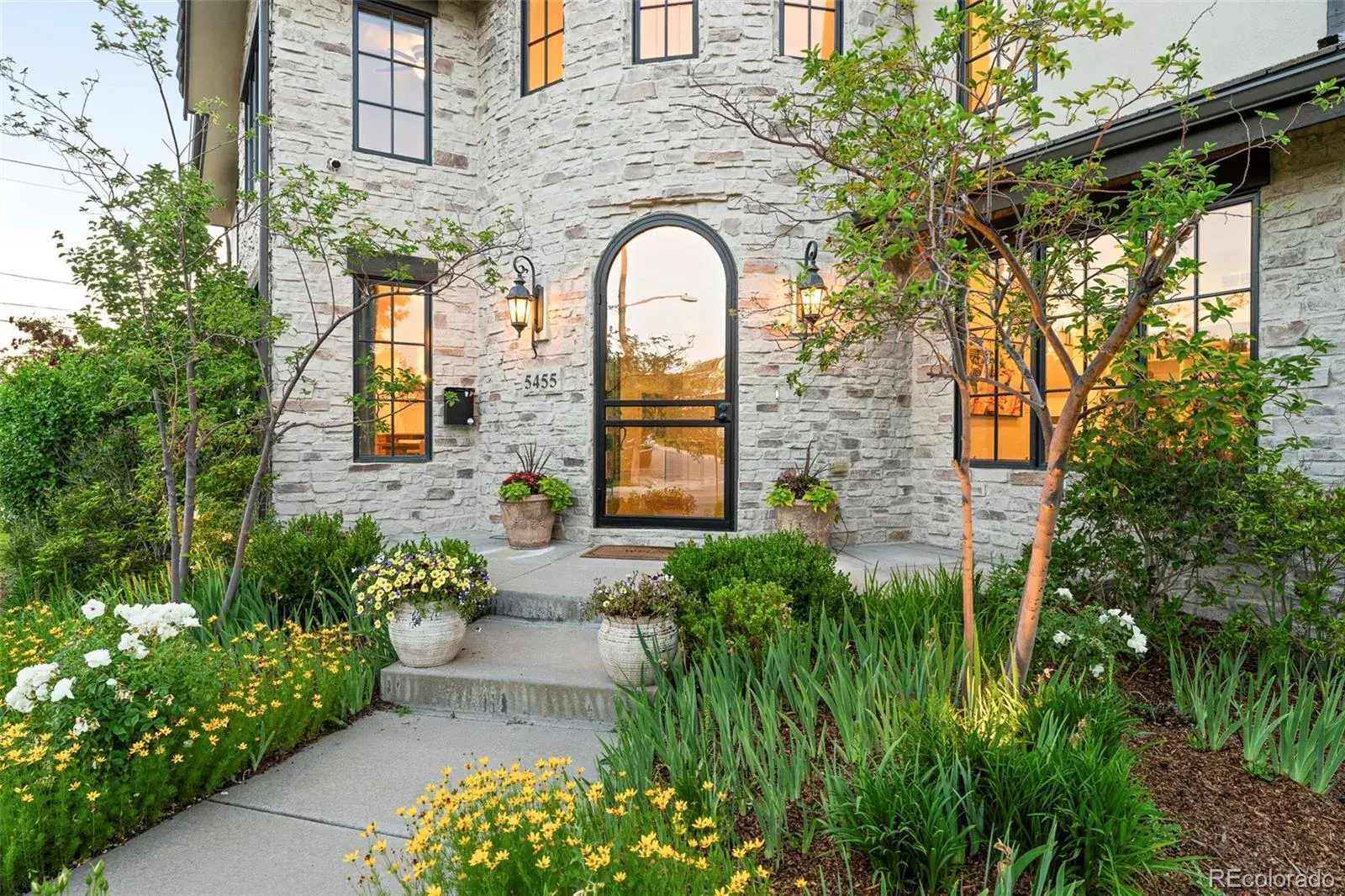 This is a front exterior view of a stone-clad house with a prominent arched entryway. The landscaping is well-maintained with a variety of flowers and greenery, adding to the curb appeal. The windows reflect the warm light of the setting sun, creating an inviting atmosphere.