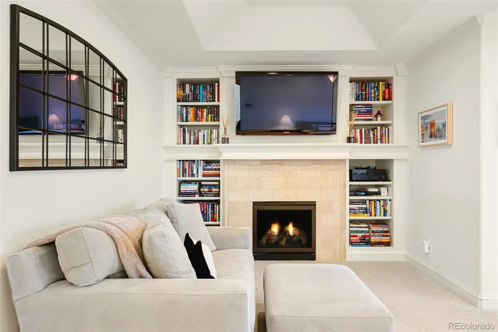 This is an inviting living room featuring a cozy fireplace with built-in bookshelves on either side, creating a focal point. A comfortable-looking sectional sofa and ottoman are arranged in front of the fireplace, suggesting a relaxing space. A large, arched window-style mirror adds architectural interest to the room.