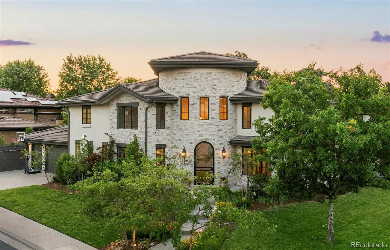This is a front exterior view of a luxurious two-story home with a unique architectural design. The house features a combination of stone and stucco, a rounded central tower, and a well-manicured lawn with mature trees and landscaping. The overall impression is one of elegance and curb appeal.