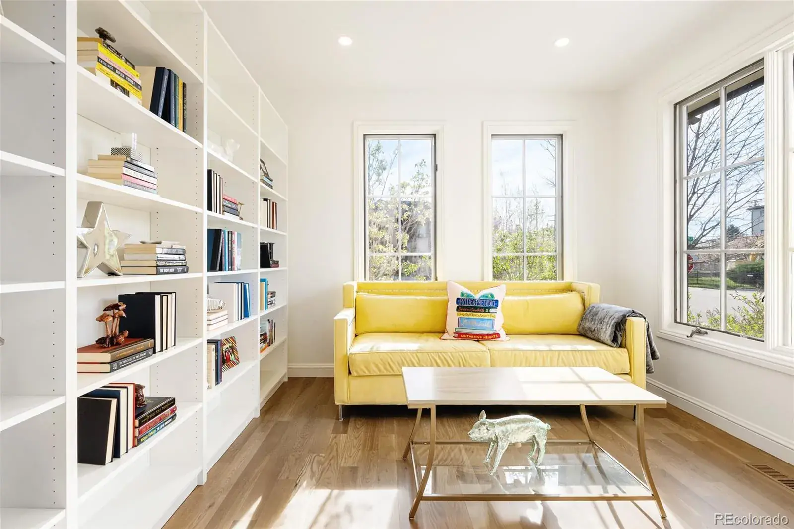 This is a bright and inviting living room featuring a large built-in bookshelf filled with books and decorative items. A yellow sofa sits beneath two windows, complemented by a modern coffee table with a glass shelf and a decorative animal figurine. The room has hardwood floors and white walls, creating a clean and airy atmosphere.